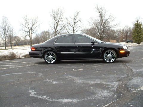 1993 Ford Taurus - Side view of a black sedan with chrome wheels parked on wet asphalt in a lot.