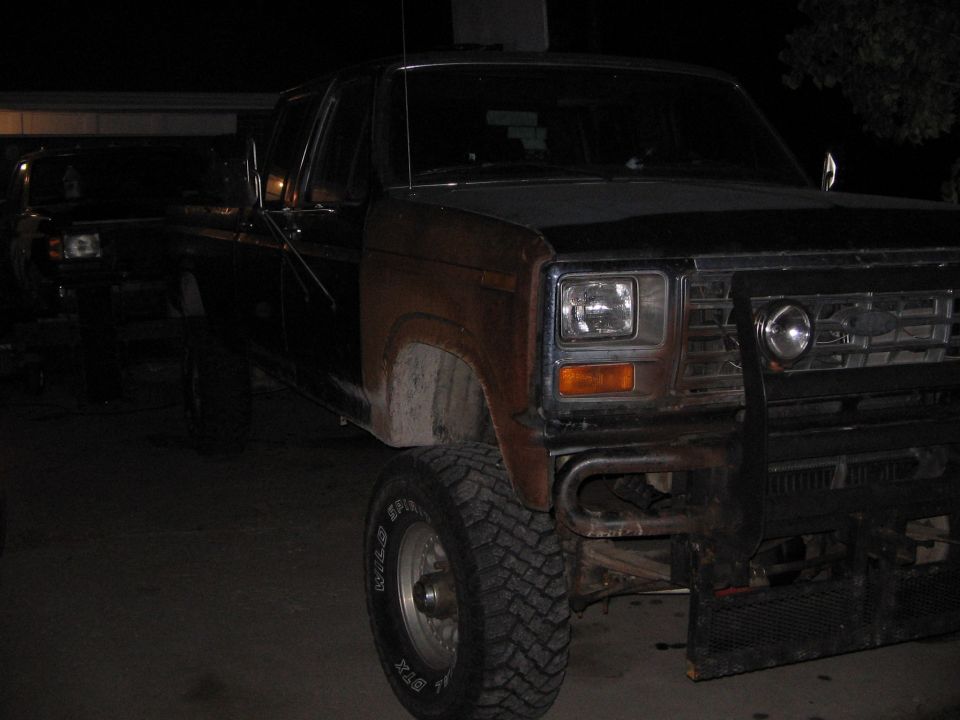 1980s Ford pickup truck with aftermarket bumper and rust on fender. Another truck is visible behind it