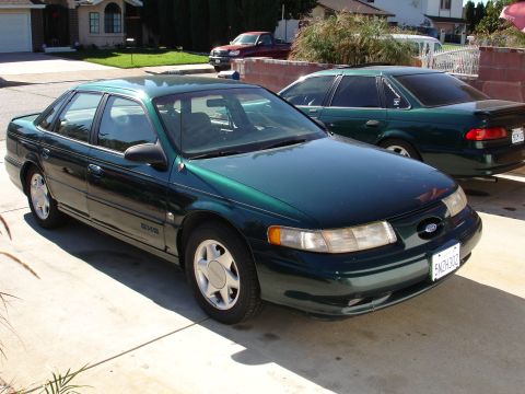 1993 Ford Taurus - A green sedan is parked in a driveway next to another car, with a house in the background.