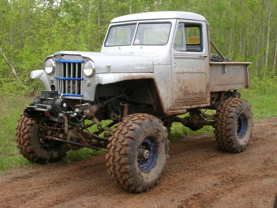 Modified Jeep pickup truck with large off-road tires and blue grille accents, covered in mud
