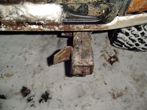 1994 Ford Bronco - Close-up of a heavily rusted metal bracket on the underside of a vehicle, with a tire and snow visible.