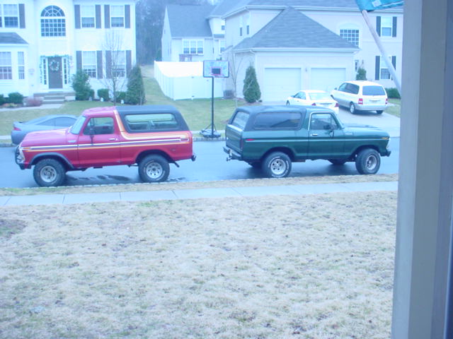 Red striped Ford Bronco parked ahead of a dark green Bronco on a suburban street, side view