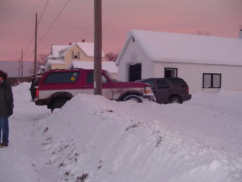1994 Ford Bronco - Two vehicles, a red and white Ford Bronco and a dark SUV, are stuck in deep snow near a house.