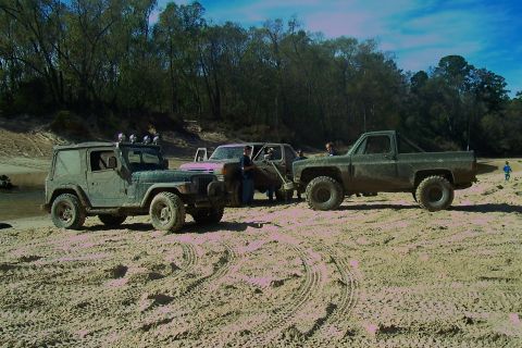 1987 Ford Bronco - A collection of off-road vehicles gathered on a sandy area near a wooded shoreline.