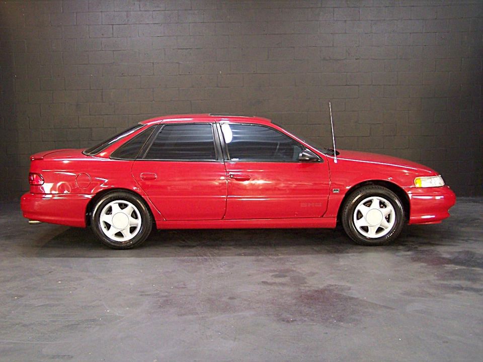 Red Ford Taurus SHO sedan, right side view, tinted windows, alloy wheels, dark block wall background