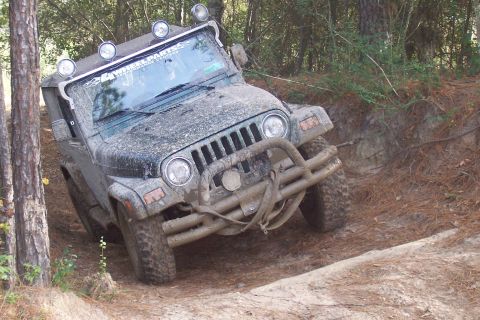 1987 Ford Bronco - A Jeep Wrangler navigates a muddy off-road trail, its body covered in dirt and equipped with auxiliary lights.