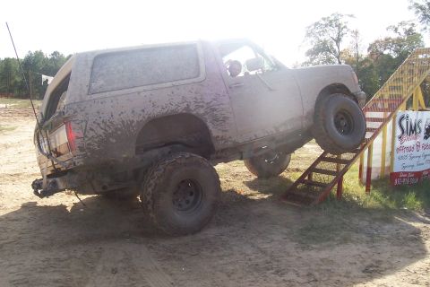 1987 Ford Bronco - A muddy SUV navigates an off-road obstacle course ramp.