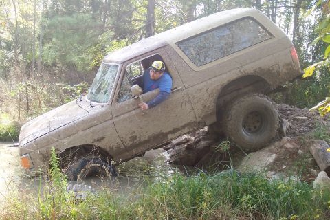 1987 Ford Bronco - A muddy SUV navigates a challenging off-road course with water and rocks.