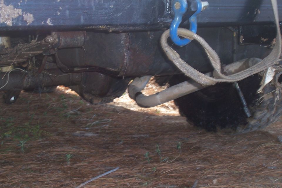 Underside of a dirty vehicle, showing suspension and exhaust pipe, with a tow strap attached to a blue shackle