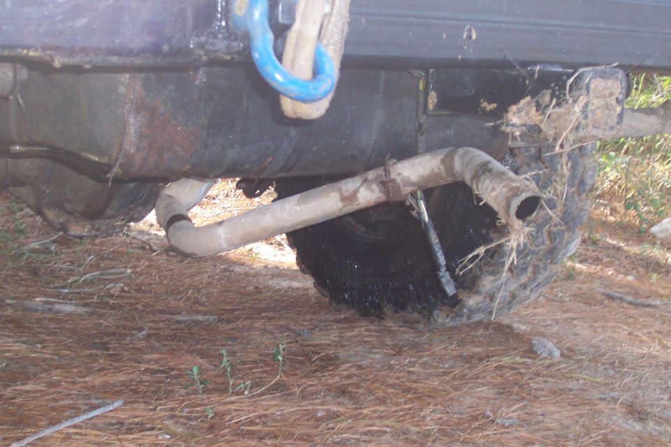 Vehicle's rear axle, exhaust pipe, and tire covered in mud, with a blue tow hook attached to the frame