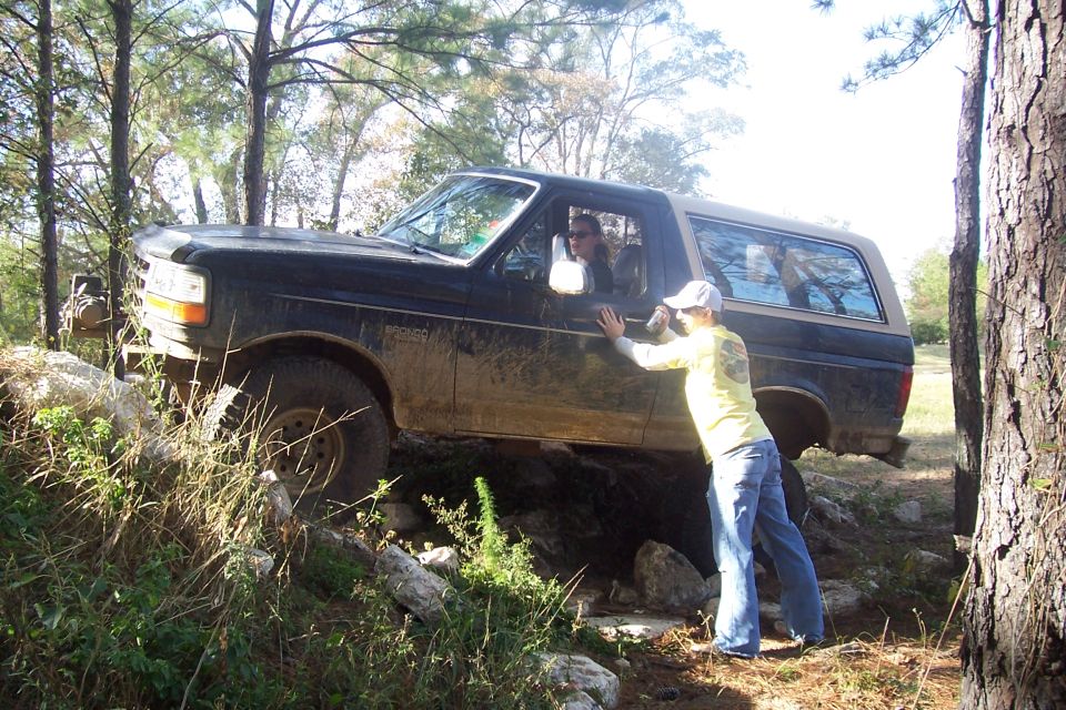 Guy pushing a black Ford Bronco on a rocky trail, driver visible inside