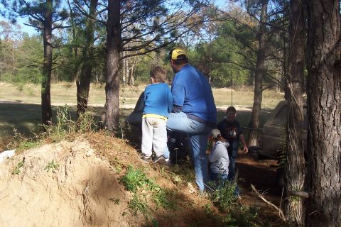 1987 Ford Bronco - A group of people are gathered in a wooded area near a dirt mound and a tent.