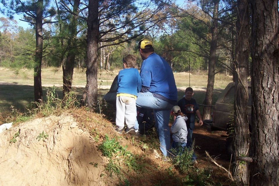 People stand around a dirt mound in a wooded area. A tent is visible behind them
