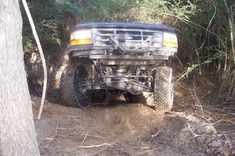1987 Ford Bronco - A black Ford Bronco navigates a muddy, wooded trail from a low front angle.
