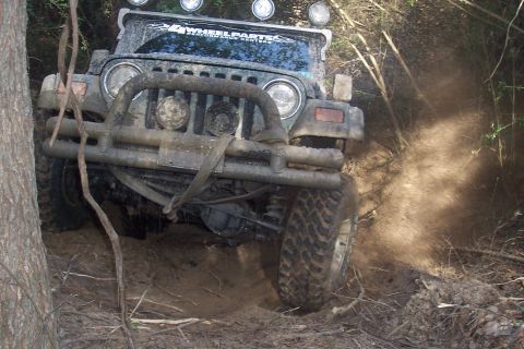 1987 Ford Bronco - Jeep Wrangler navigating a muddy, wooded trail with dust kicking up.