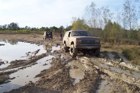 1987 Ford Bronco - A 1987 Ford Bronco is pulled through a muddy terrain by a Jeep, with trees and a body of water in the background.