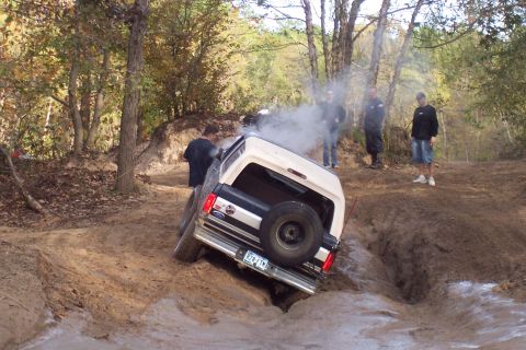 1987 Ford Bronco - A 1987 Ford Bronco is stuck in a muddy ditch, emitting smoke, with several people watching.