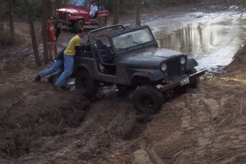 1987 Ford Bronco - Two Jeeps off-roading in mud, one requires assistance from bystanders.