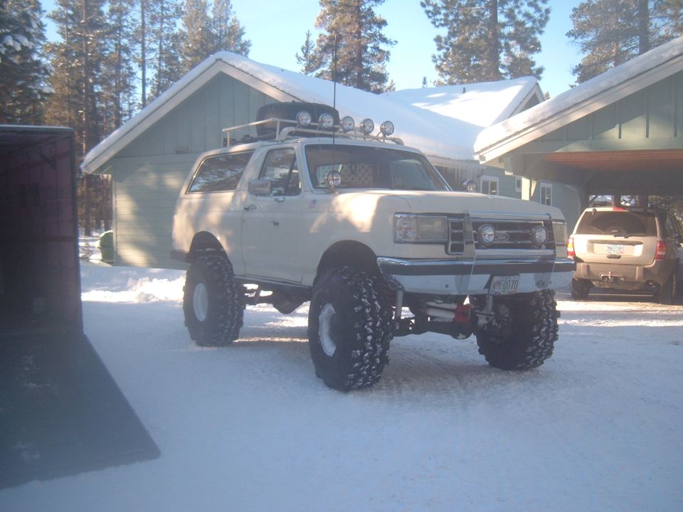 Lifted white Ford Bronco on mud tires with roof rack and light bar, parked in snow