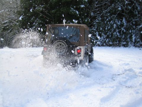 1990 Ford Bronco - A Jeep Wrangler drives through deep snow, sending spray into the air.