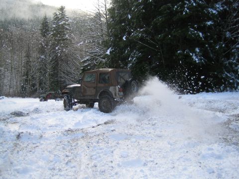 1990 Ford Bronco - A Jeep Wrangler drives through snow, spraying it up as it goes. Another Jeep is visible in the distance.