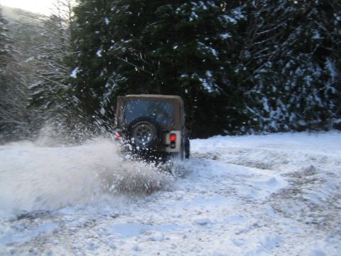 1990 Ford Bronco - A Jeep Wrangler navigates a snowy path, sending snow flying as it turns.