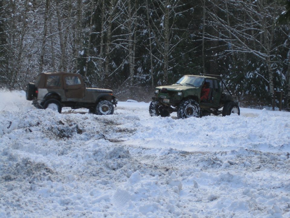 Two modified off-road vehicles driving through deep snow in a wooded area