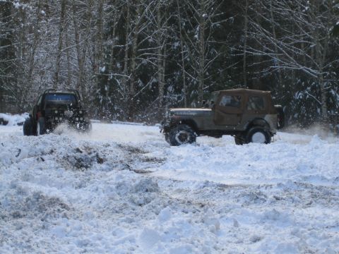 1990 Ford Bronco - Two off-road vehicles navigate a snowy, wooded terrain, kicking up snow as they drive.