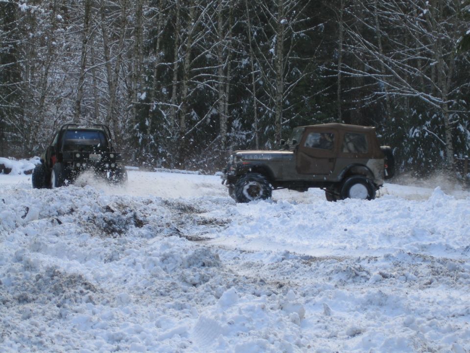 Two off-road vehicles driving through deep snow in a wooded area. One is a dark SUV, the other a tan Jeep