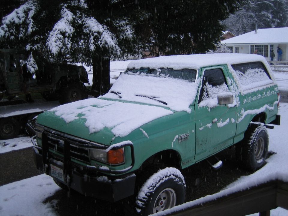 Green 1990 Ford Bronco covered in snow, parked next to another vehicle