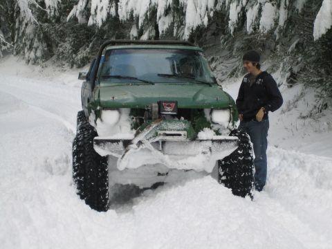 1990 Ford Bronco - A person stands beside a modified green SUV partially buried in snow on a wooded road.