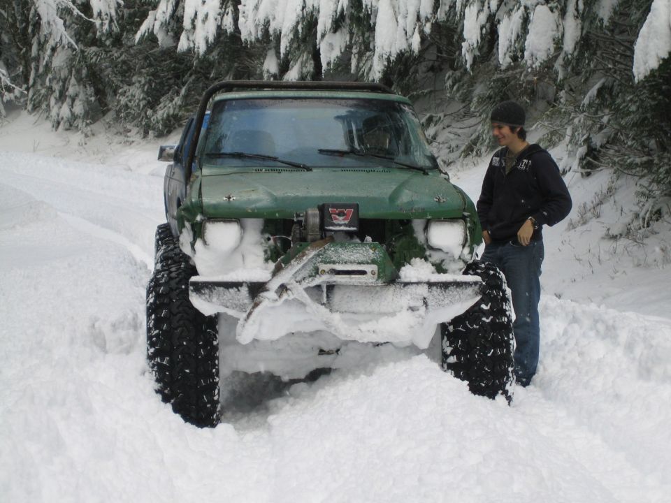 Green SUV with large tires stuck in snow, front bumper removed, engine exposed