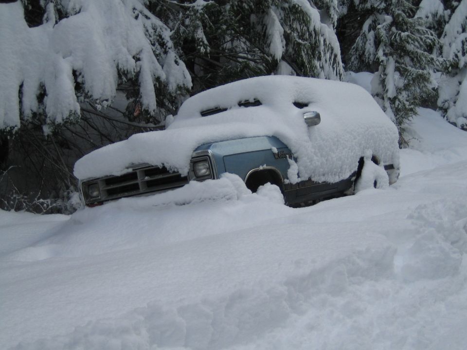 Light blue pickup truck, possibly 1980s Dodge Ram, covered in deep snow with trees in background