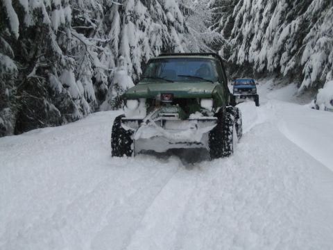 1990 Ford Bronco - Two off-road vehicles navigate a snow-covered forest road, with the lead vehicle equipped with a snow plow.