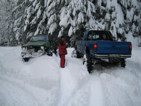 1990 Ford Bronco - Two modified trucks are stuck in a snowy forest, with one vehicle showing a 'Chevrolet' badge on its tailgate.