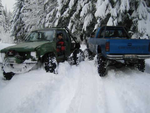 1990 Ford Bronco - Two modified trucks are stuck in a snowy forest, with one showing significant front-end damage.