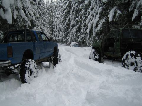 1990 Ford Bronco - Two modified trucks navigate a snow-covered forest road, kicking up powder.