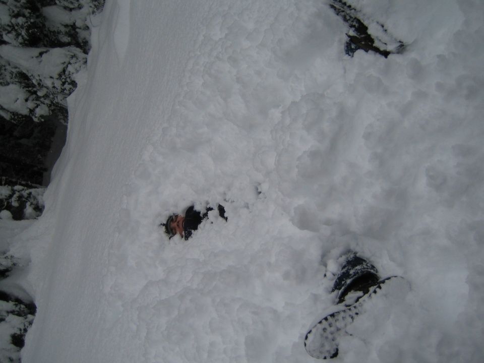 Person partially buried in snow, with boots visible. A rock face is to the left