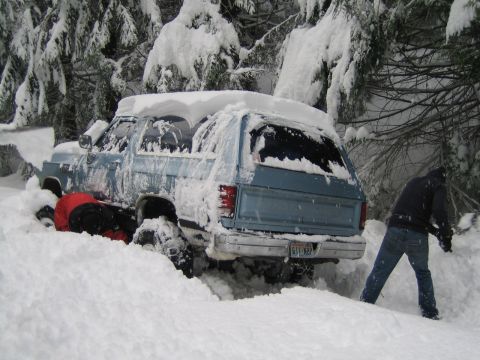 1990 Ford Bronco - A person digs out a blue SUV that is stuck deep in the snow, surrounded by snow-covered trees.
