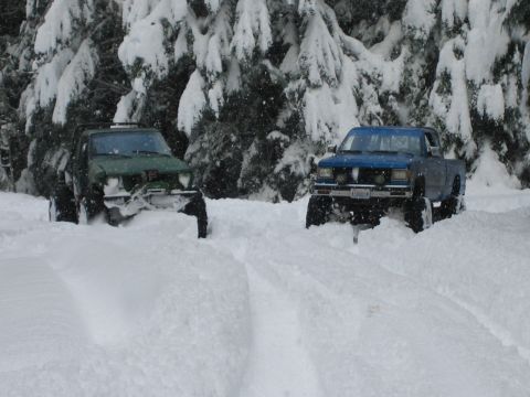 1990 Ford Bronco - Two modified trucks navigate a snowy forest road, kicking up powder as they go.