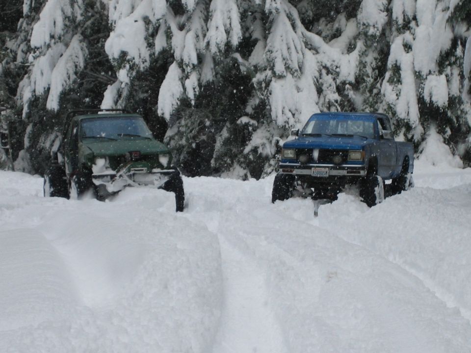 Blue pickup truck with large tires driving through deep snow, green off-road vehicle behind it