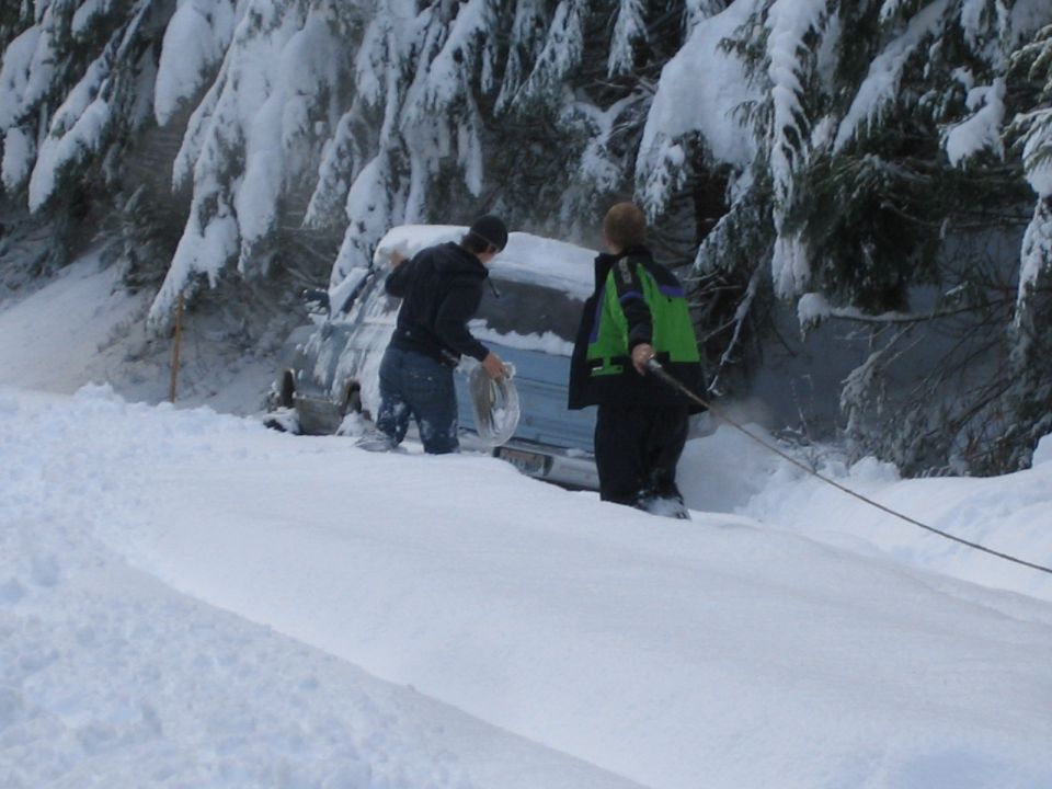 Two people are trying to pull a vehicle stuck in deep snow next to a wooded area