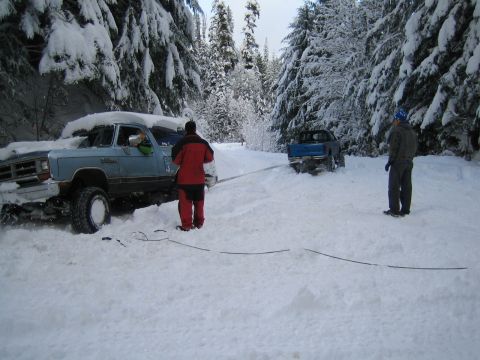 1990 Ford Bronco - Two trucks are stuck in the snow on a wooded road, with one attempting to tow the other.