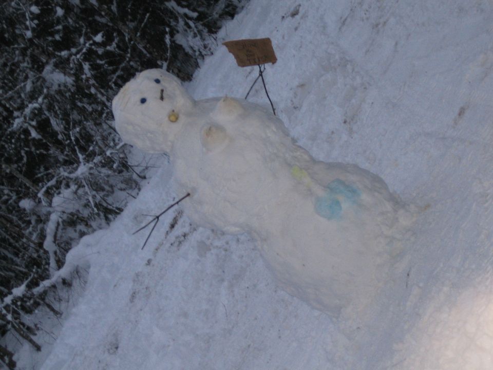 Snowman lying down in snow with stick arms and a small sign