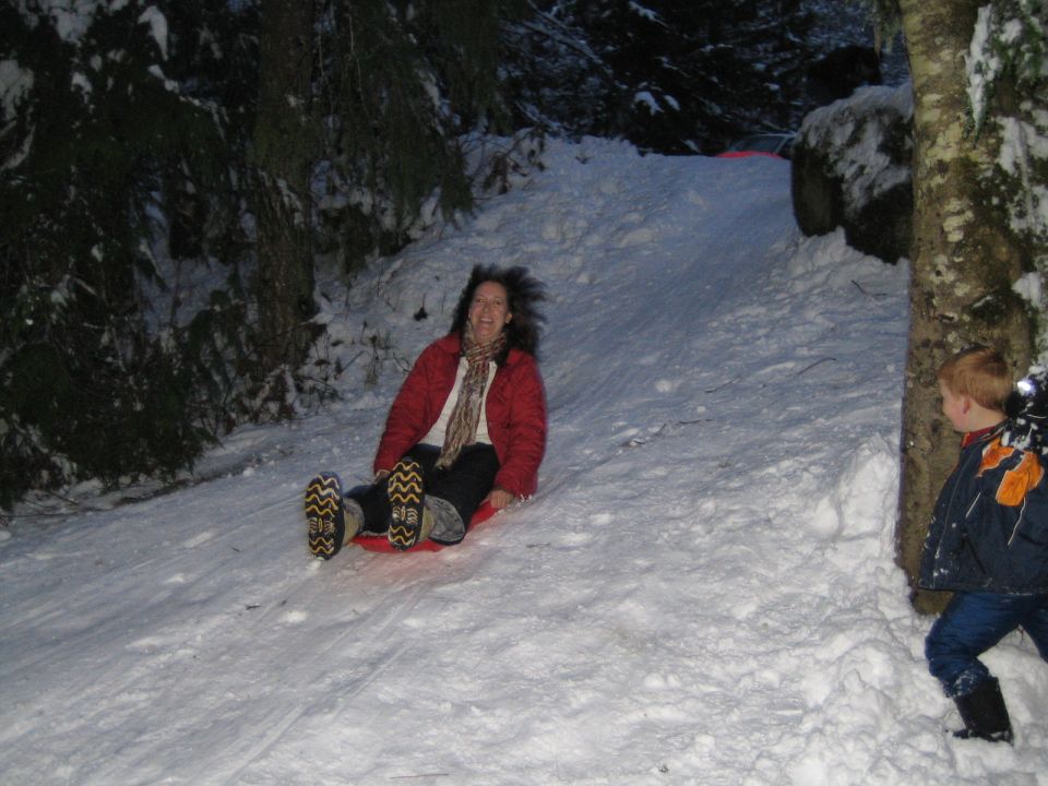 Woman on a red sled going down a snow-covered hill, with a young boy standing to the right