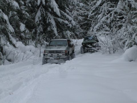 1990 Ford Bronco - Two 4x4 vehicles navigate a snow-covered trail through a dense, winter forest.