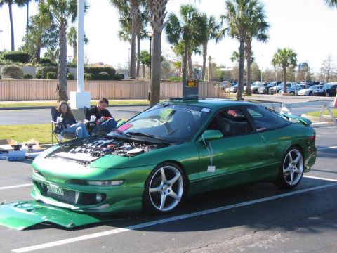 1994 Ford Probe - A green coupe with its hood and bumper removed sits in a parking lot, attended by two people in lawn chairs.