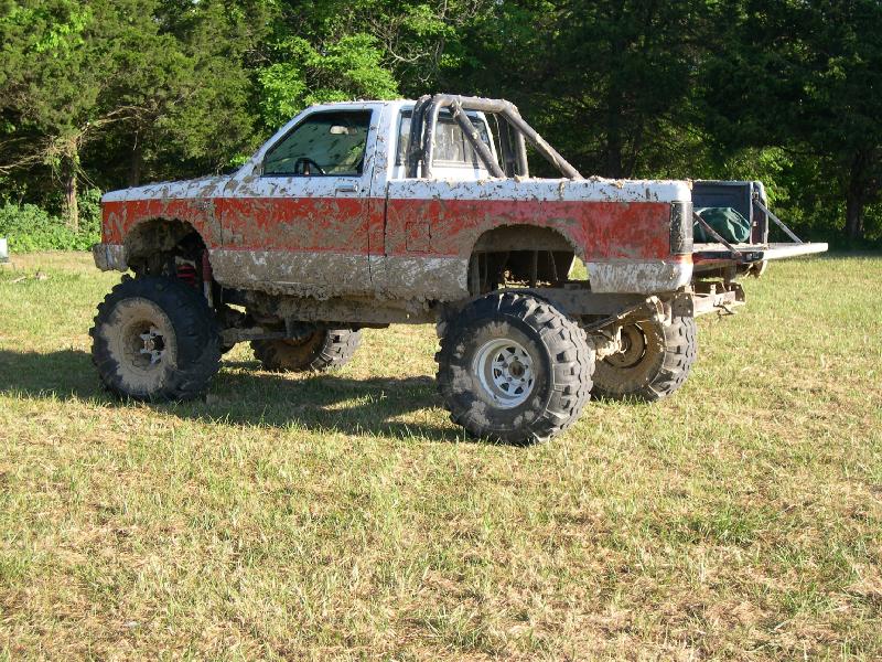 Mud-covered red and white lifted mini pickup with roll bar and big mud tires in a field