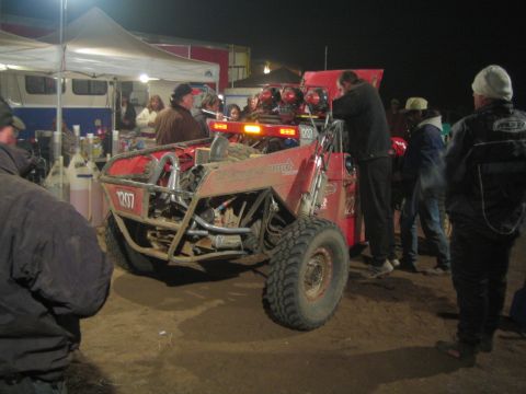 1990 Ford Bronco - red number-1207 off-road buggy sits in a brightly lit pit area surrounded by tents and crew at night.