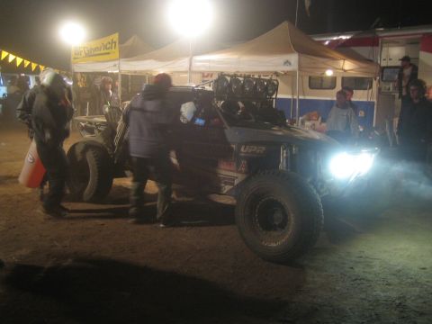 1990 Ford Bronco - off-road race buggy with bright headlights sits among canopy tents and crew in a busy nighttime pit.